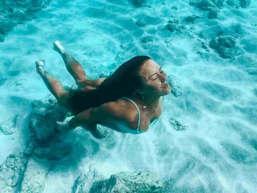 Donna nuota sott'acqua, superando la paura delle acque profonde. La luce filtra attraverso l'acqua cristallina, illuminando la sabbia e le rocce sul fondale.