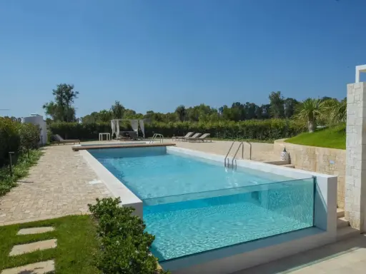 Piscina fuori terra su terreno in pendenza con pareti in vetro, circondata da un giardino curato e un'area relax con lettini e gazebo.