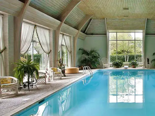 Piscina interna con acqua azzurra, sedie in vimini, piante e grandi finestre. Uno skimmer piscina cos'è? Serve a raccogliere detriti galleggianti.