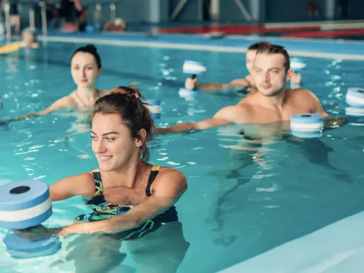 Persone sorridenti fanno sport in piscina con attrezzi galleggianti per un allenamento acquatico tonificante.
