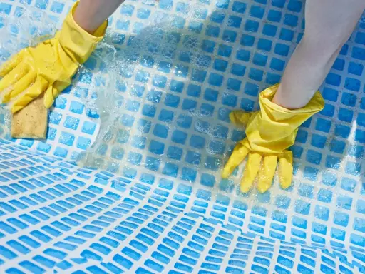 Mani con guanti gialli puliscono il fondo di una piscina con una spugna, mostrando come togliere calcare dalla piscina.