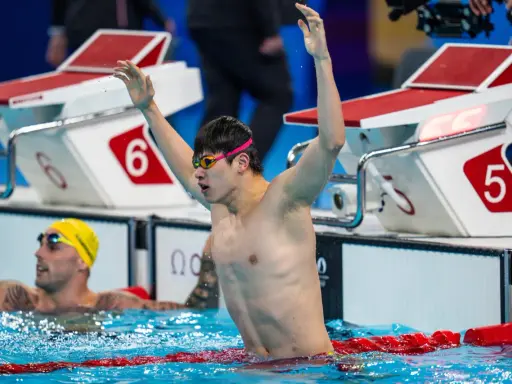 Nuotatore con braccia alzate, festeggia la vittoria. Pan Zhanle trionfa in piscina, circondato da avversari e blocchi di partenza.