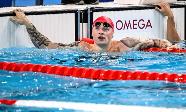 Adam Peaty, con cuffia rossa e occhialini, emerge dall'acqua della piscina, aggrappato al bordo.