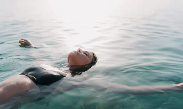 Donna galleggia sull'acqua, superando la paura di nuotare dove non si tocca. La luce del sole illumina il suo viso sereno.