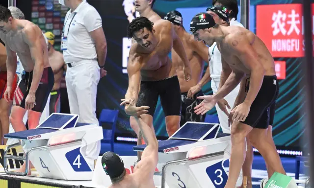 Atleti italiani festeggiano dopo la staffetta 4x100. Un nuotatore porge la mano a un compagno, gioia e sudore bagnano la corsia.