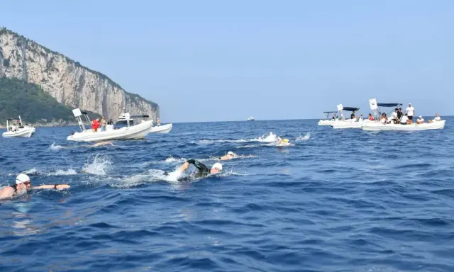 Nuotatori in gara tra Capri e Napoli, con barche di supporto e la costa rocciosa sullo sfondo.