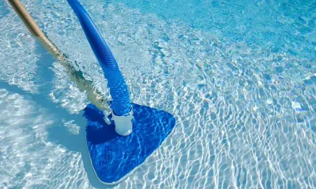 Pulizia di una piscina sporca con aspiratore blu e manico in legno. L'acqua limpida riflette la luce creando un effetto scintillante.