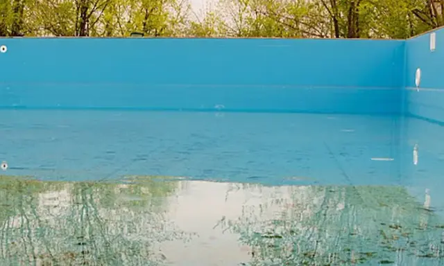La piscina in inverno va svuotata. L'acqua residua riflette il verde degli alberi e il cielo, mentre il fondo mostra i segni del tempo.