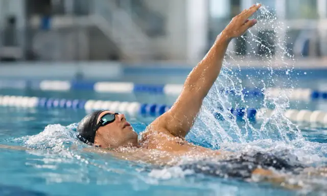 Atleta in piscina esegue la nuotata a **scheda dorso**, con il braccio destro sollevato e l'acqua che schizza.