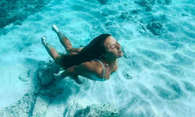 Donna nuota sott'acqua, superando la paura delle acque profonde. La luce filtra attraverso l'acqua cristallina, illuminando la sabbia e le rocce sul fondale.