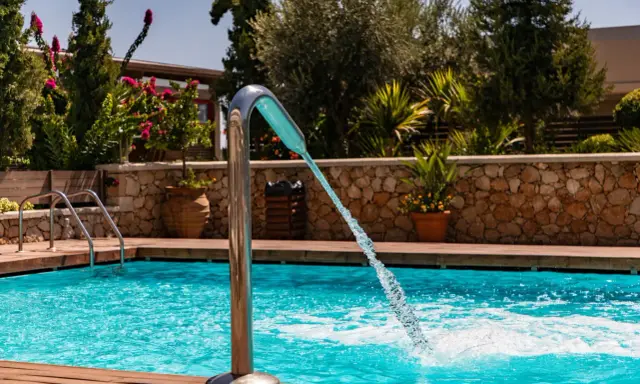 Un getto d'acqua da una fontana metallica riempie la piscina azzurra, circondata da legno e vegetazione lussureggiante.