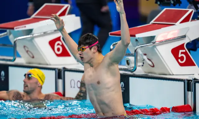 Nuotatore con braccia alzate, festeggia la vittoria. Pan Zhanle trionfa in piscina, circondato da avversari e blocchi di partenza.