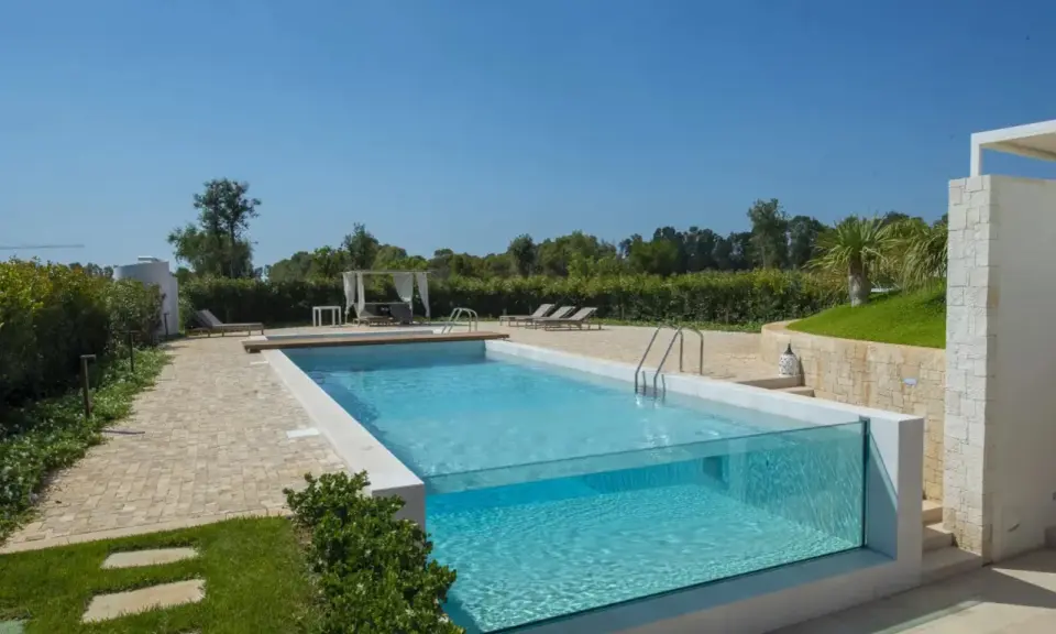 Piscina fuori terra su terreno in pendenza con pareti in vetro, circondata da un giardino curato e un'area relax con lettini e gazebo.