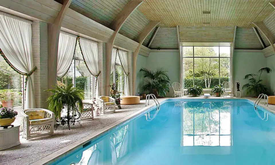 Piscina interna con acqua azzurra, sedie in vimini, piante e grandi finestre. Uno skimmer piscina cos'è? Serve a raccogliere detriti galleggianti.