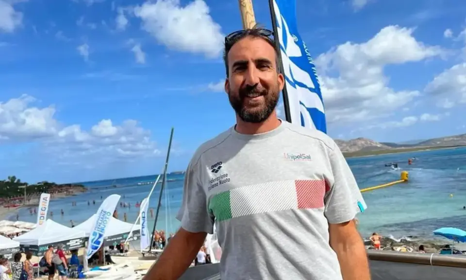 Fabrizio Antonelli sorride in spiaggia, con la bandiera italiana sul petto, pronto per un evento di nuoto.