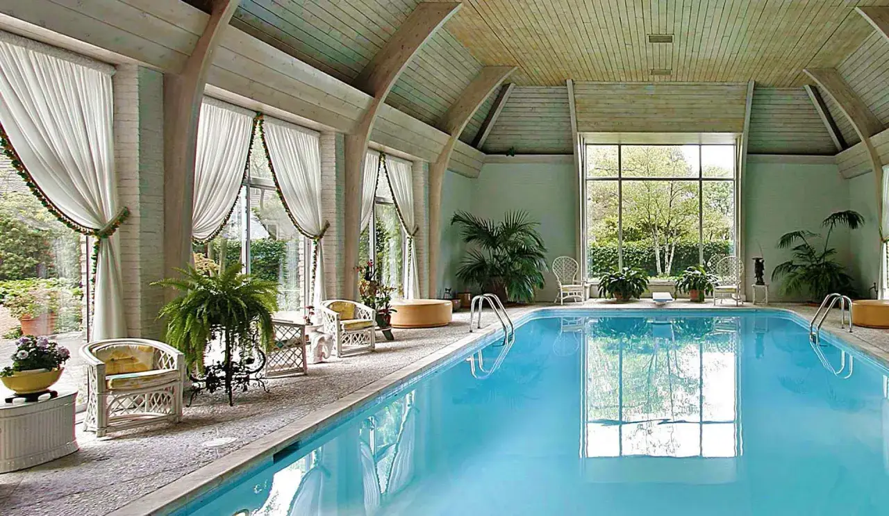 Piscina interna con acqua azzurra, sedie in vimini, piante e grandi finestre. Uno skimmer piscina cos'è? Serve a raccogliere detriti galleggianti.