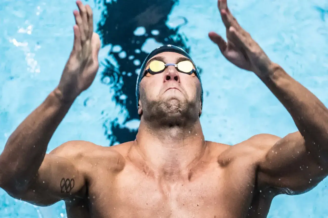 Matt Grevers, nuotatore con occhialini e cuffia, alza le braccia dall'acqua blu, mostrando la sua determinazione.