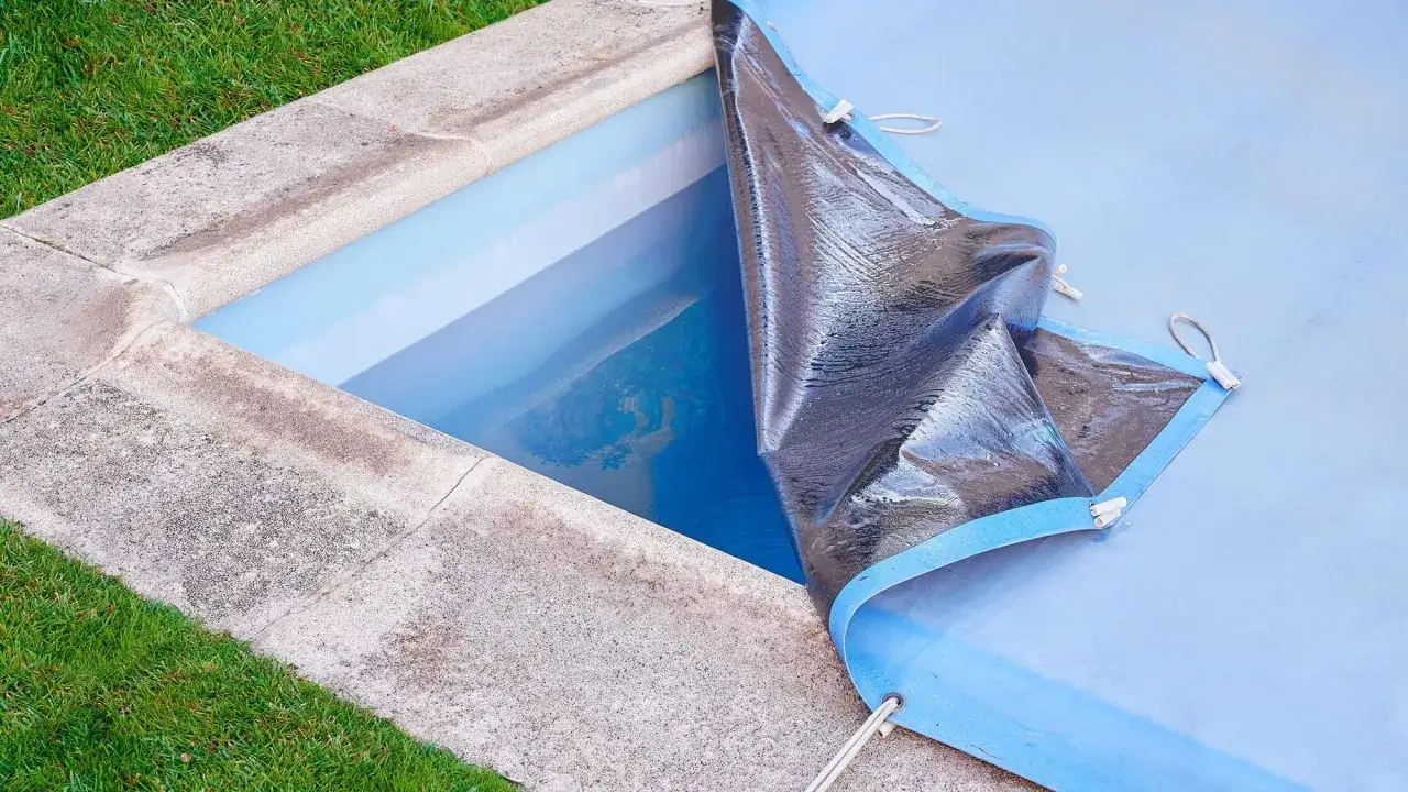 Piscina con telo in PVC parzialmente sollevato, pronto per la pulizia. L'acqua azzurra riflette il cielo.