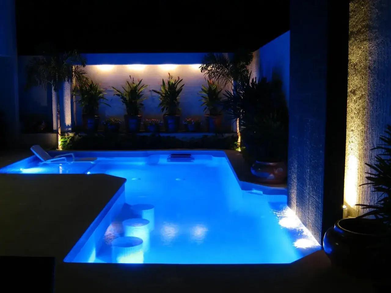 Una splendida piscina di sera, illuminata da luci blu e gialle, con piante tropicali e una cascata d'acqua.