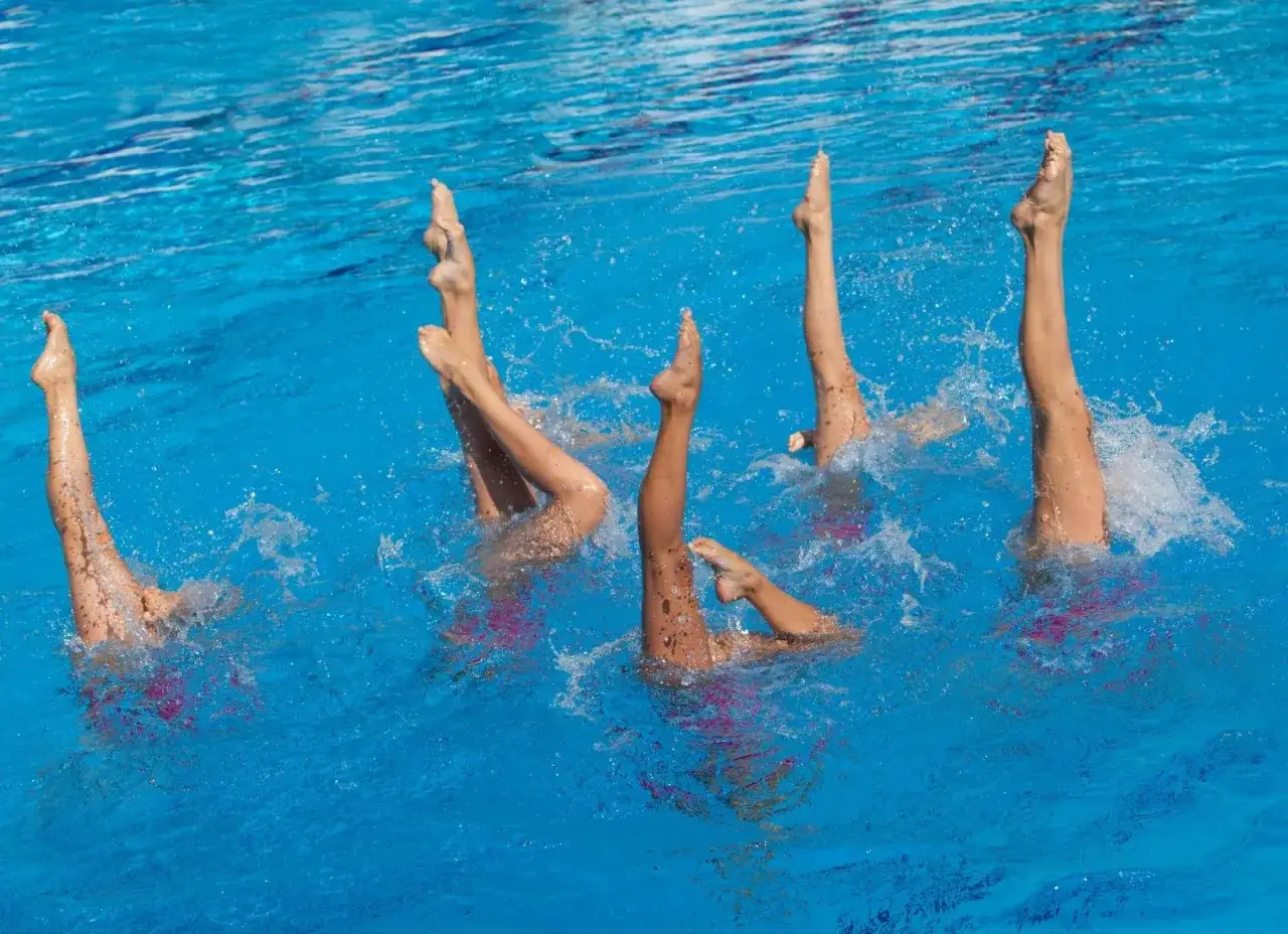 Atlete di nuoto sincronizzato eseguono una figura con le gambe in aria, circondate da spruzzi d'acqua.