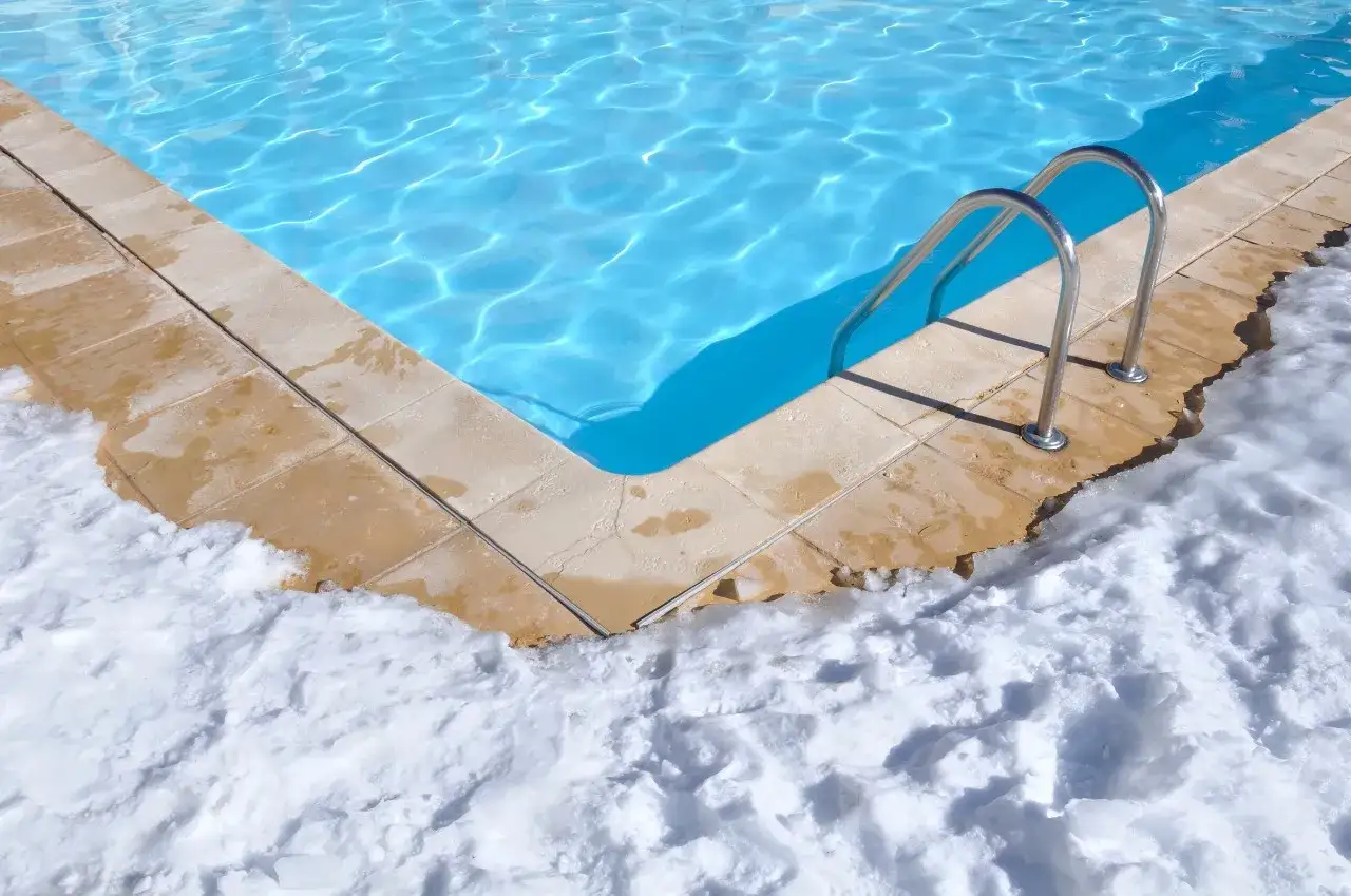 Angolo di una piscina con acqua azzurra e bordo piastrellato, circondato da neve. La piscina in inverno va svuotata per proteggerla dal gelo.