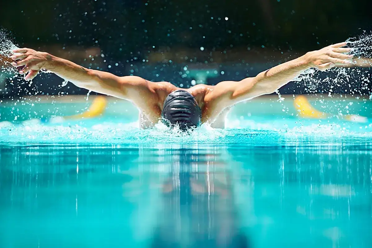 Atleta in pieno stile farfalla, con un movimento di stretching che precede il nuoto. L'acqua schizza intorno a lui.