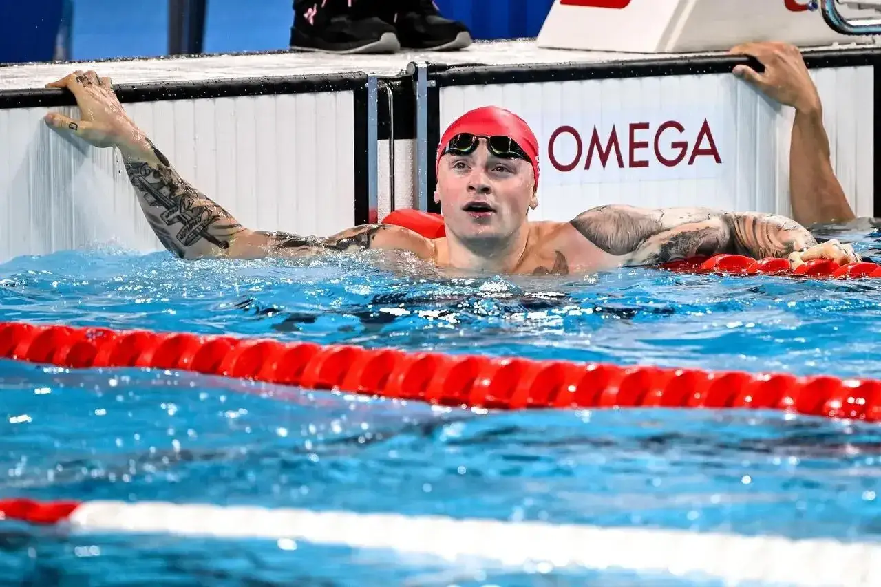 Adam Peaty, con cuffia rossa e occhialini, emerge dall'acqua, aggrappato al bordo della piscina. Tatuaggi coprono le sue braccia.